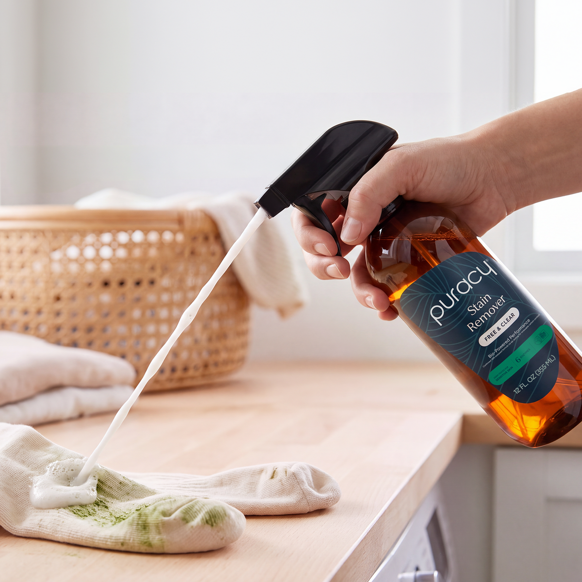 Hand sprays Laundry Stain Remover for New Customers on dirty socks; laundry basket in background.