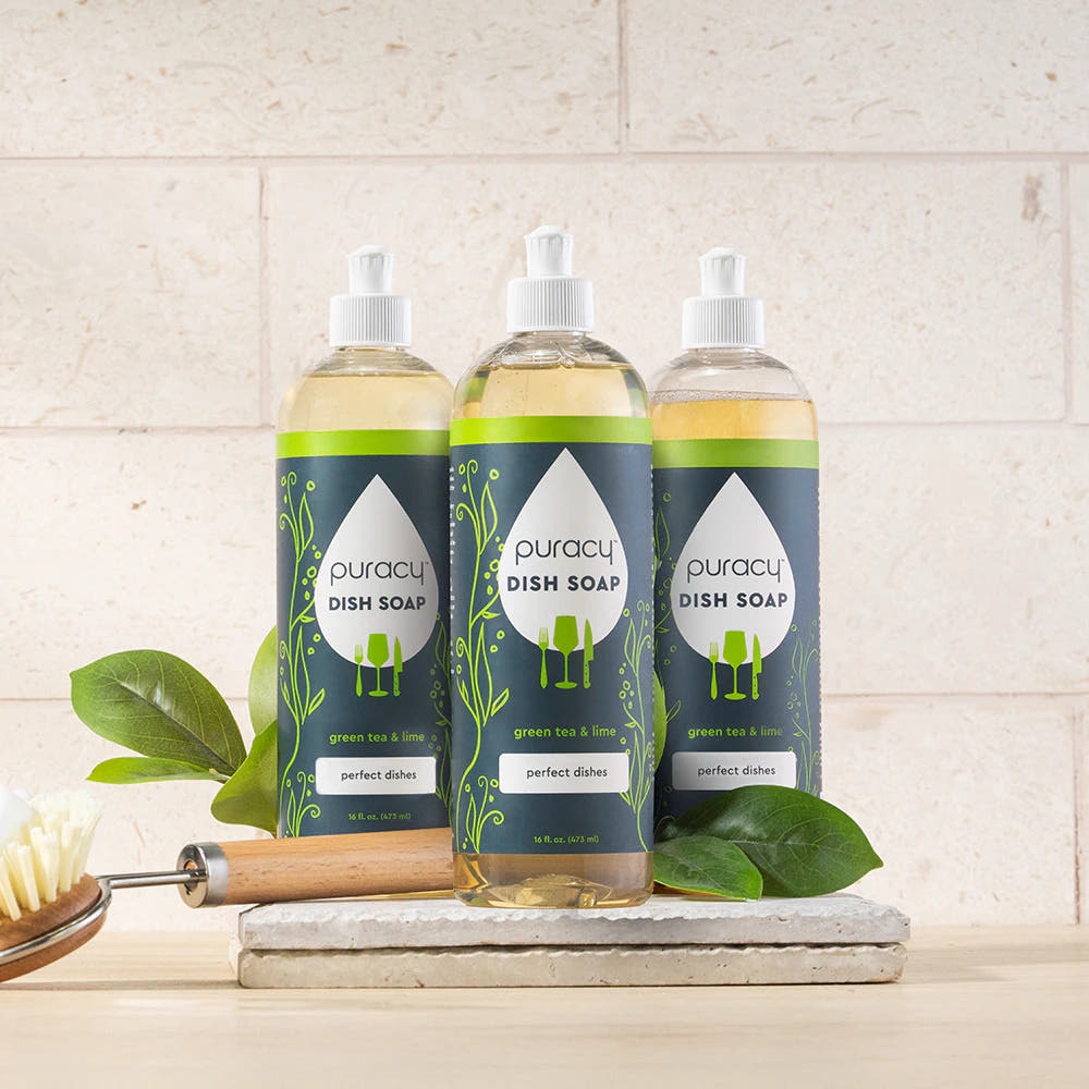 Three bottles of Puracy Dish Soap with green labels are on a counter with leaves and a brush.