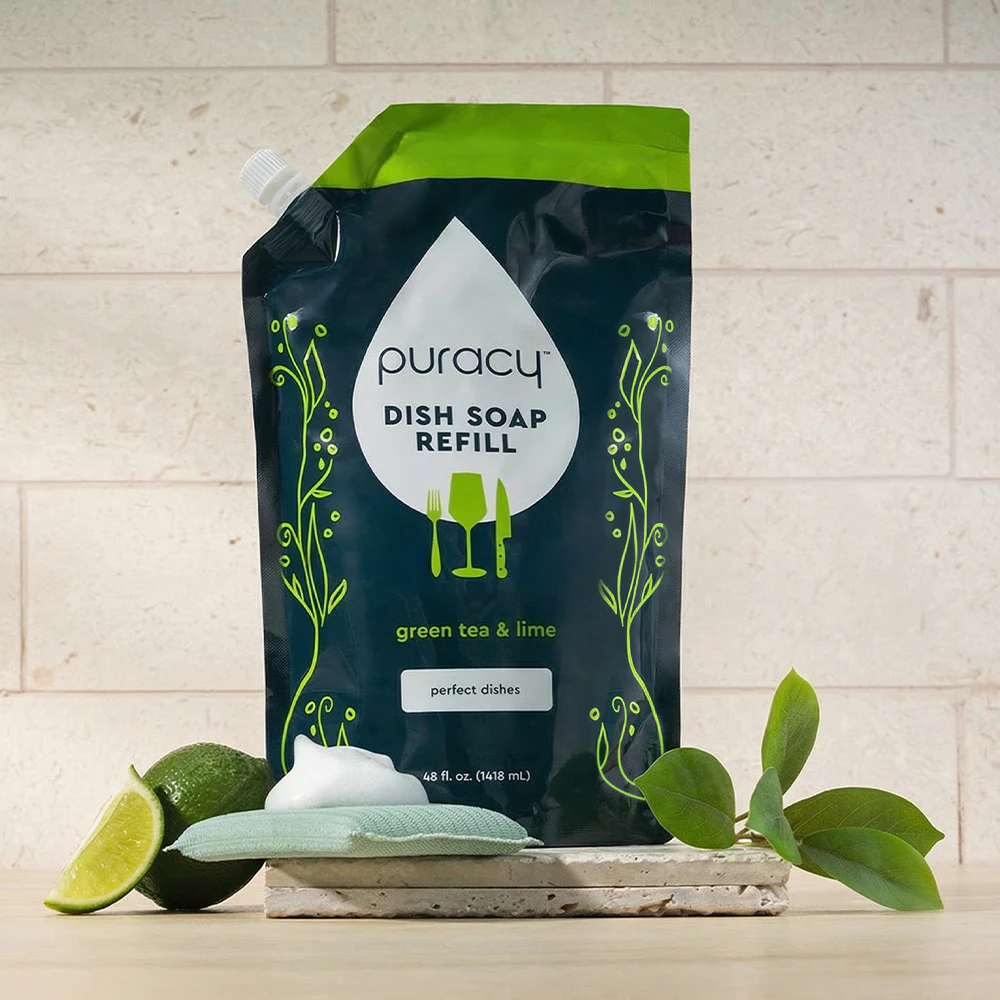 Puracy Dish Soap refill pouch with sponge, lime, and leaves on a kitchen counter.