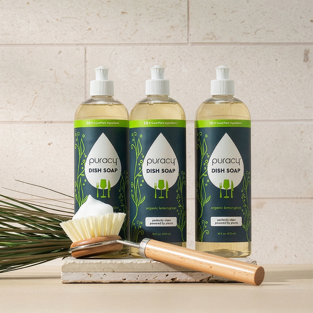 Three bottles of Puracy Dish Soap next to a cleaning brush and green plant on a countertop.