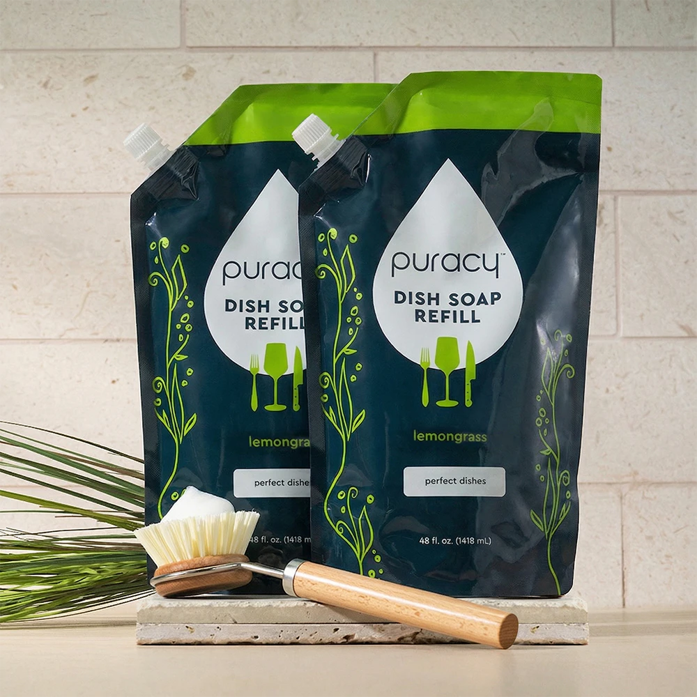 Two Puracy Dish Soap refill pouches sit by a dish brush on the kitchen counter.