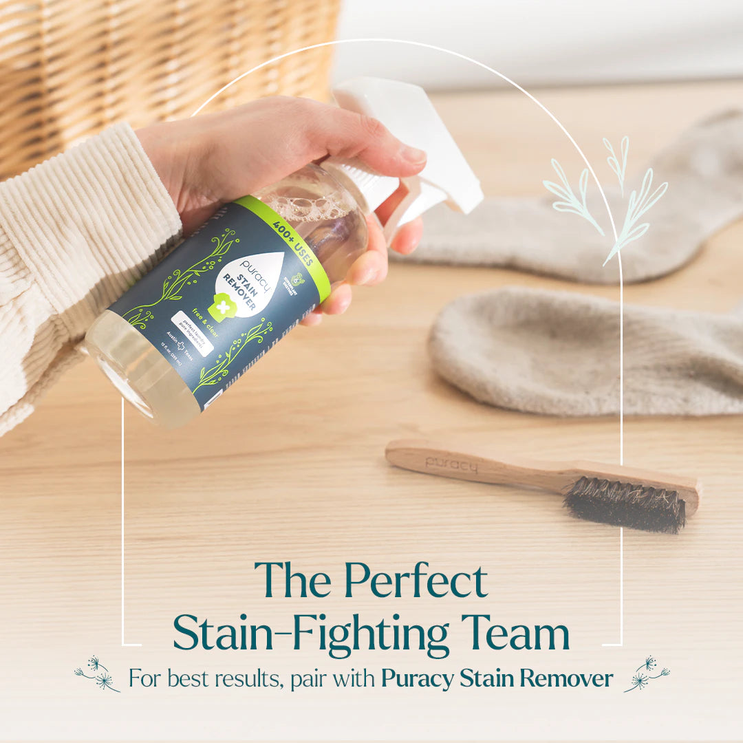 Person holding a bottle of Puracy stain remover with a brush and fabric on a wooden surface.