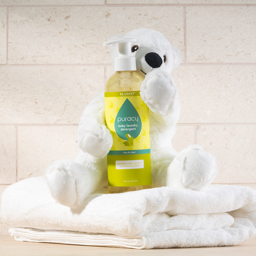 A white plush bear hugs a bottle of Baby Laundry Detergent atop neatly folded white towels.