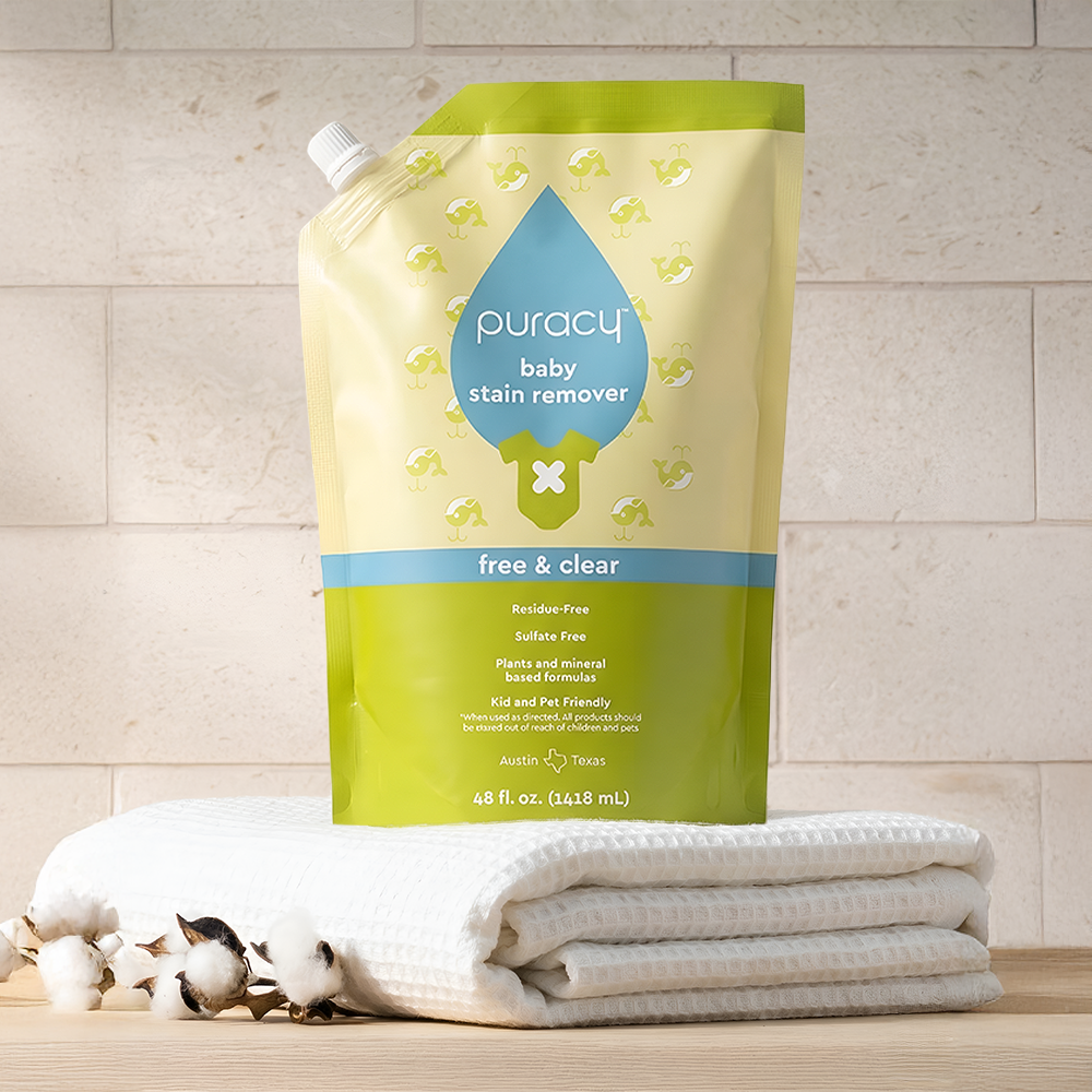 A pouch of Baby Stain Remover sits on folded towels beside cotton branches.