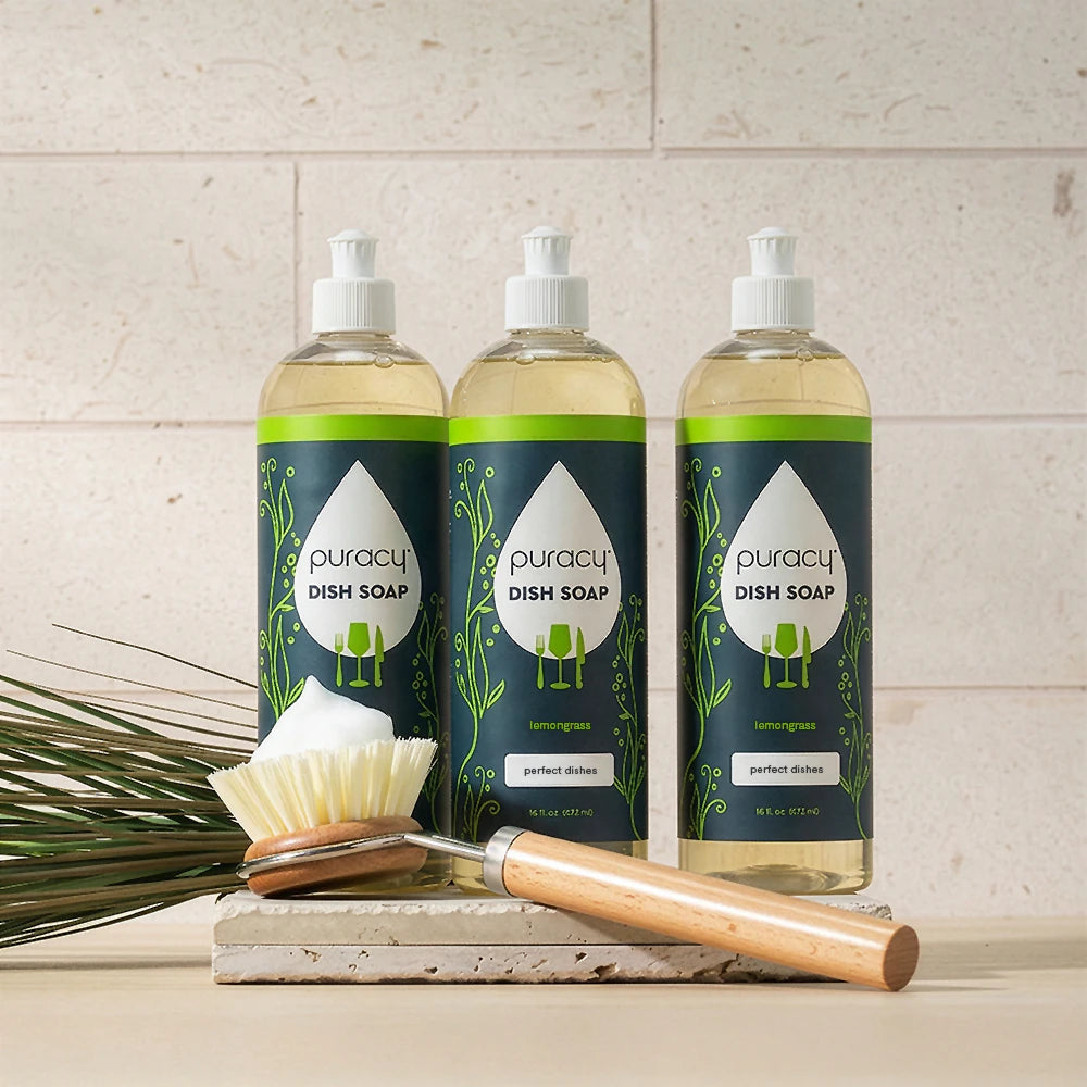 Three bottles of Dish Soap with a brush and green plant on a white surface.