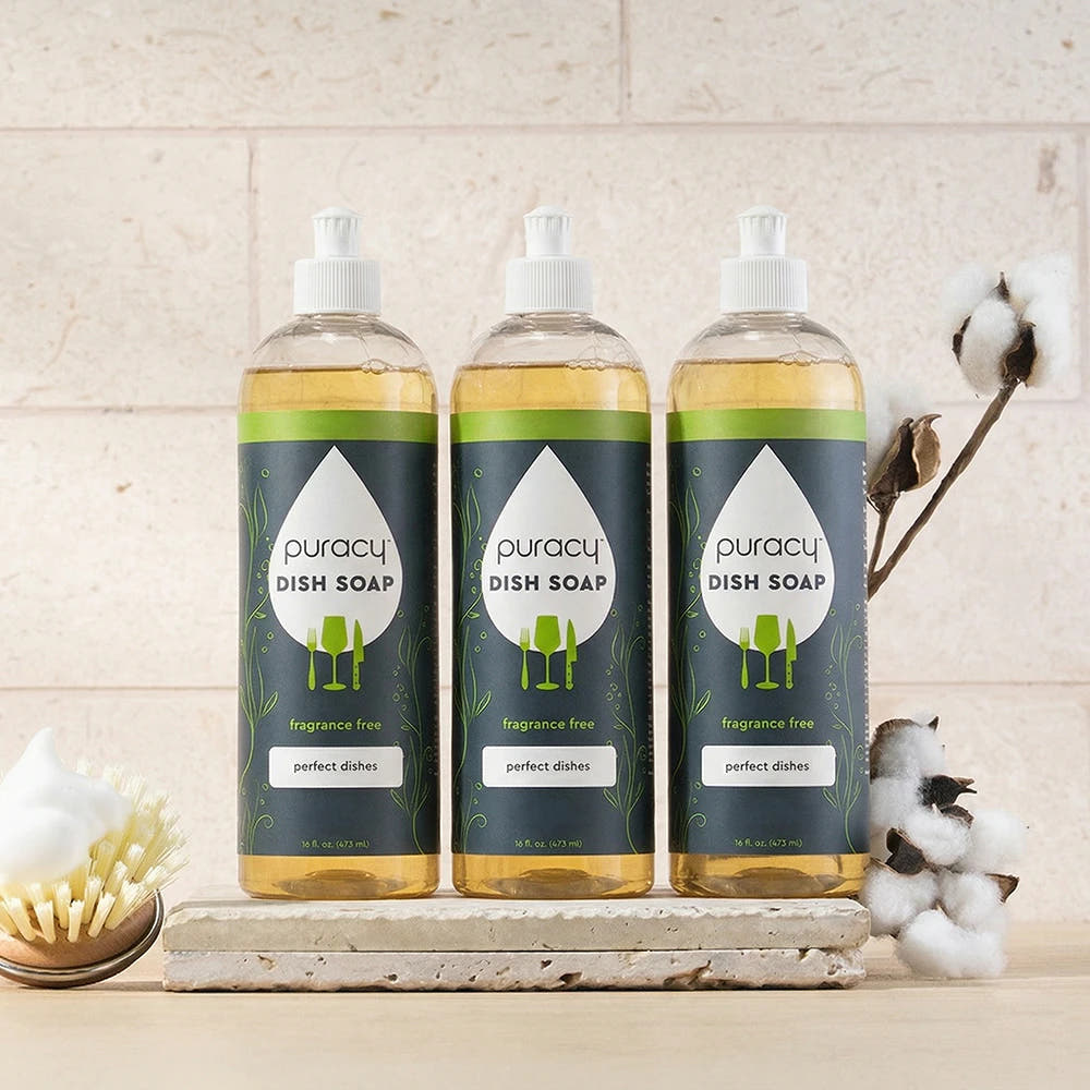 Three bottles of Puracy Dish Soap with cotton and a dish brush on a light countertop.