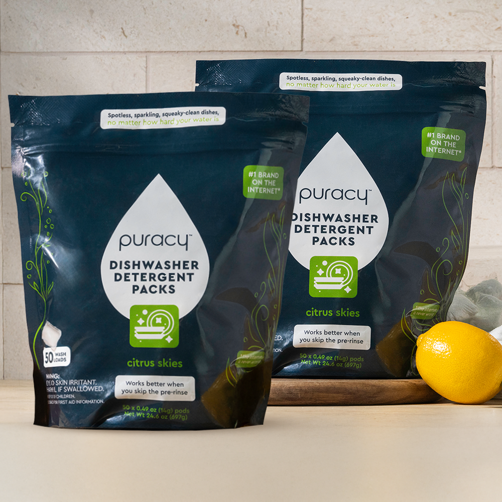 Two Dishwasher Detergent Pods packs sit on a counter with a lemon and a green towel.
