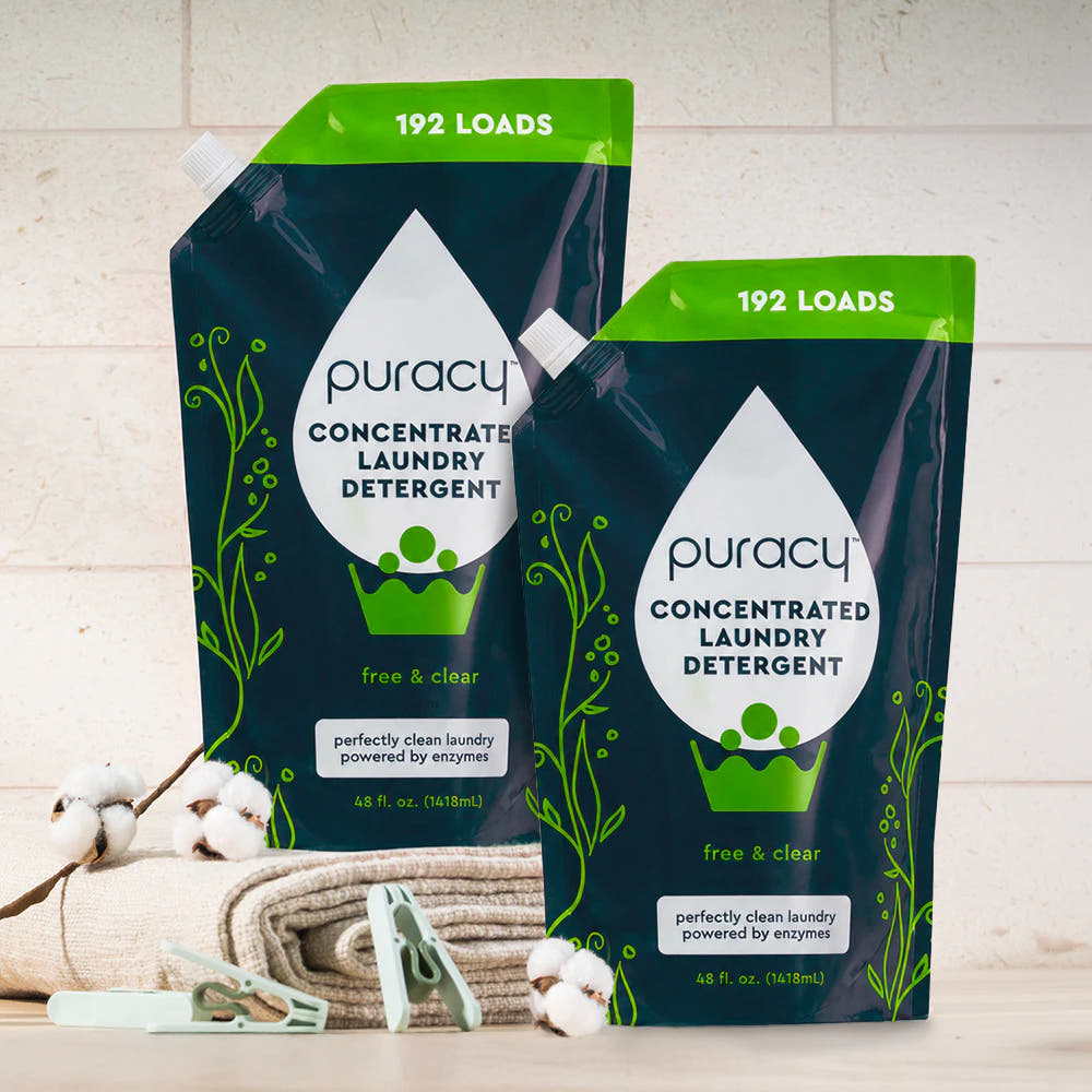 Two sulfate-free Puracy concentrated laundry detergent refill pouches beside folded towels on a counter.