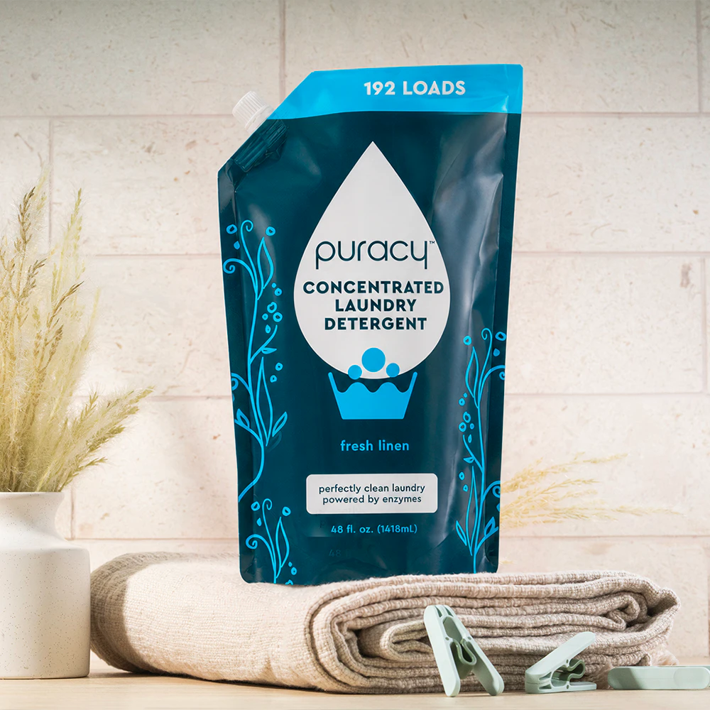 A blue pouch of Puracy Laundry Detergent (Concentrated, Sulfate-Free) sits on folded towels and clothespins.